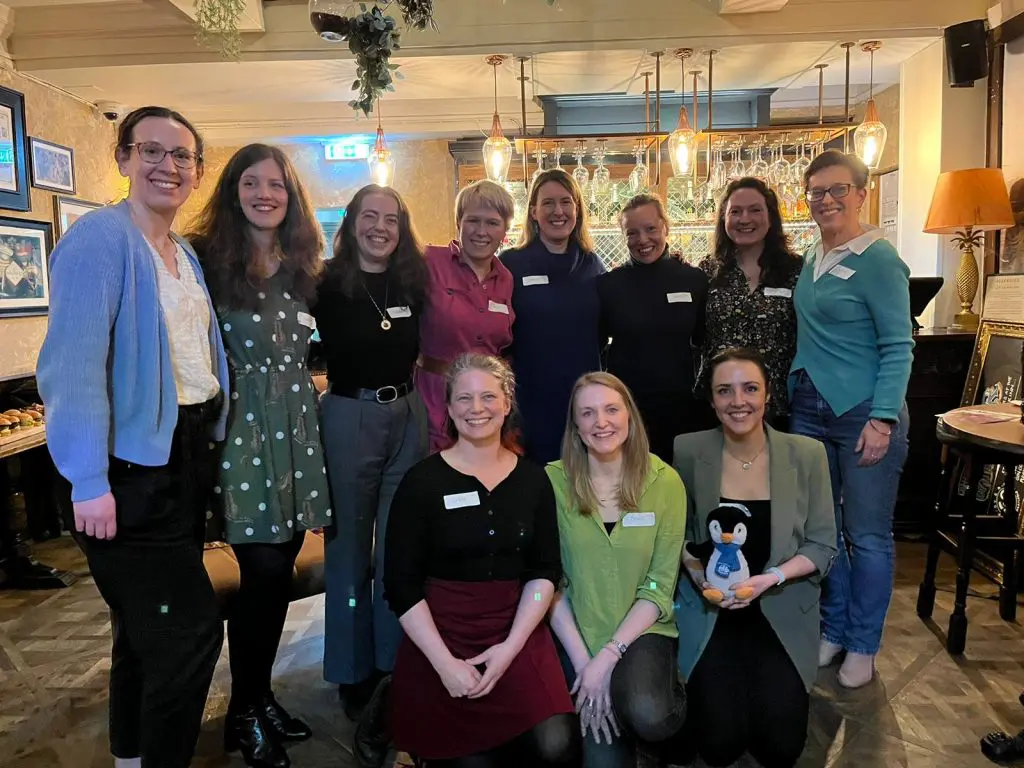 group of white women in casual dress pose, smiling, for the camera. One holds a small cuddly penguin.