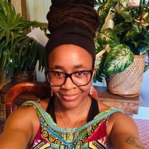 Nnedi is a black woman with large black rimmed glasses and a tattoo on her upper right arm. Her hair is tied up under a black scarf with some dreadlocks wrapped around the outside of the scarf. She is wearing a colourful tank top and is smiling at the camera
