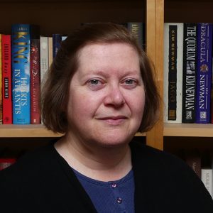 Claire is a white woman, shown from the shoulders up. Claire has short chin length brown hair and is wearing a blue top with a black cardigan. She is stood in front of shelves of books.
