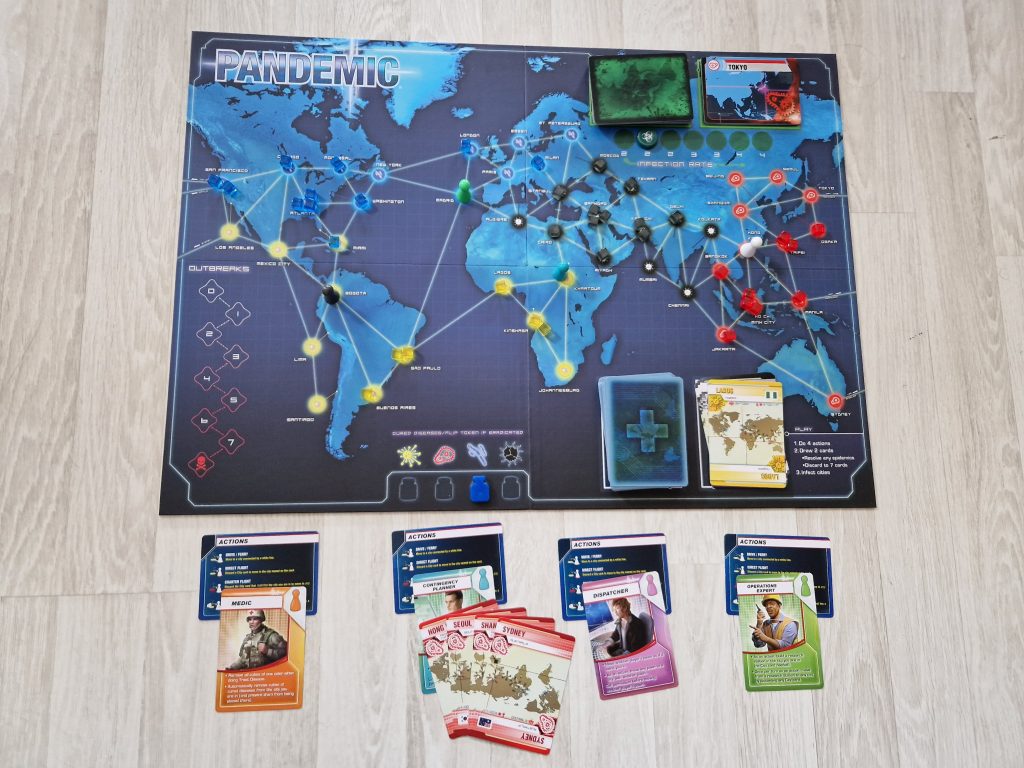 A blue game board on a light wooden floor. The game board has a blue-tinted map of six continents with colored dots with connecting white lines printed on it. Across the top of the game board are small black, red, yellow, and blue plastic game pieces and green, black, and white plastic player pieces. In the upper and lower right corners of the board, there sit two stacks of game cards. Below the board are four stacks of various drawn game play cards which feature actions, roles, and locations. 