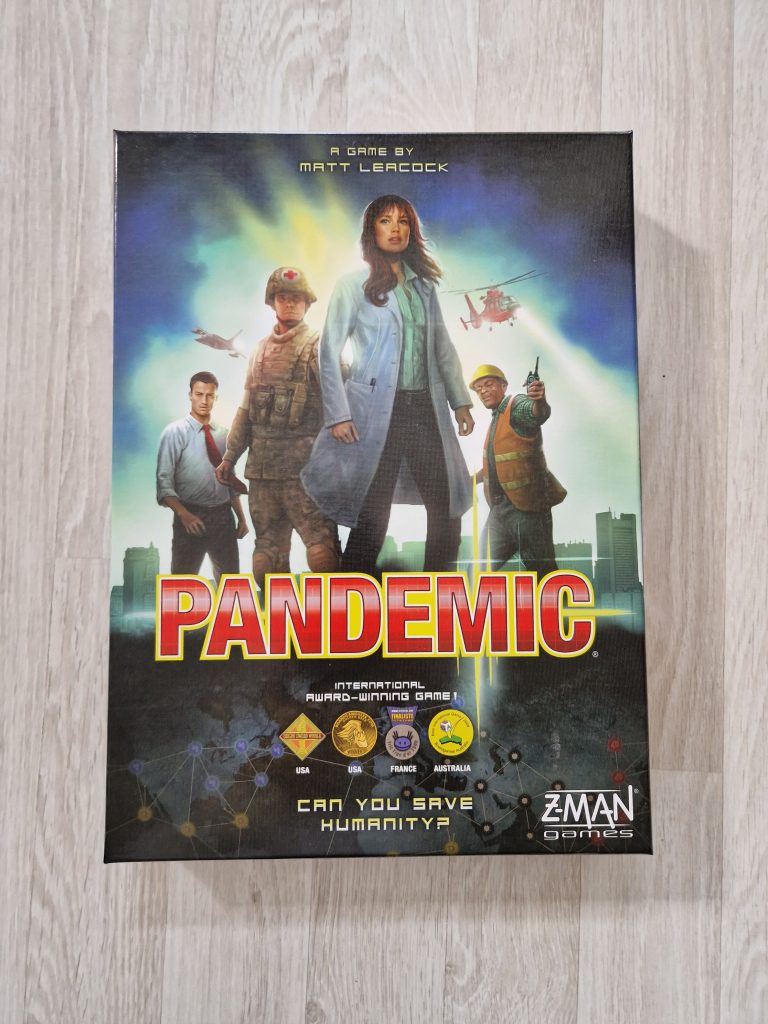 The box for the tabletop game "Pandemic" is pictured on a light wooden floor. The box art includes a doctor, medic, administrator, emergency worker, and a helicopter on a background with buildings low on the horizon and a dark blue and green clouds looming above. The title reads "Pandemic" in red block type. The slogan text "Can You Save Humanity?" appears at the bottom below four award icons. 