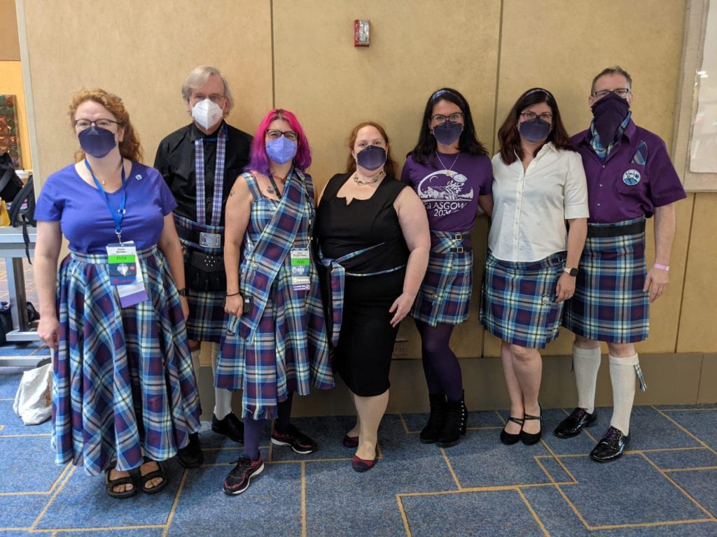 Photo shows seven people standing in a row. They are pictured wearing face masks and various garments made of Landing Zone Glasgow tartan including kilts, skirts, shawls, scarves, and sashes. 