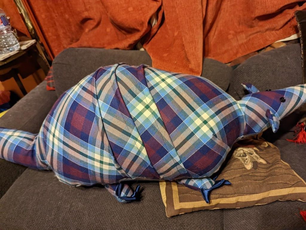 A stuffed toy armadillo made of purple, blue, green, cream, and silver tartan fabric sits on top of a brown pillow on a brown couch. The toy is pictured in profile.