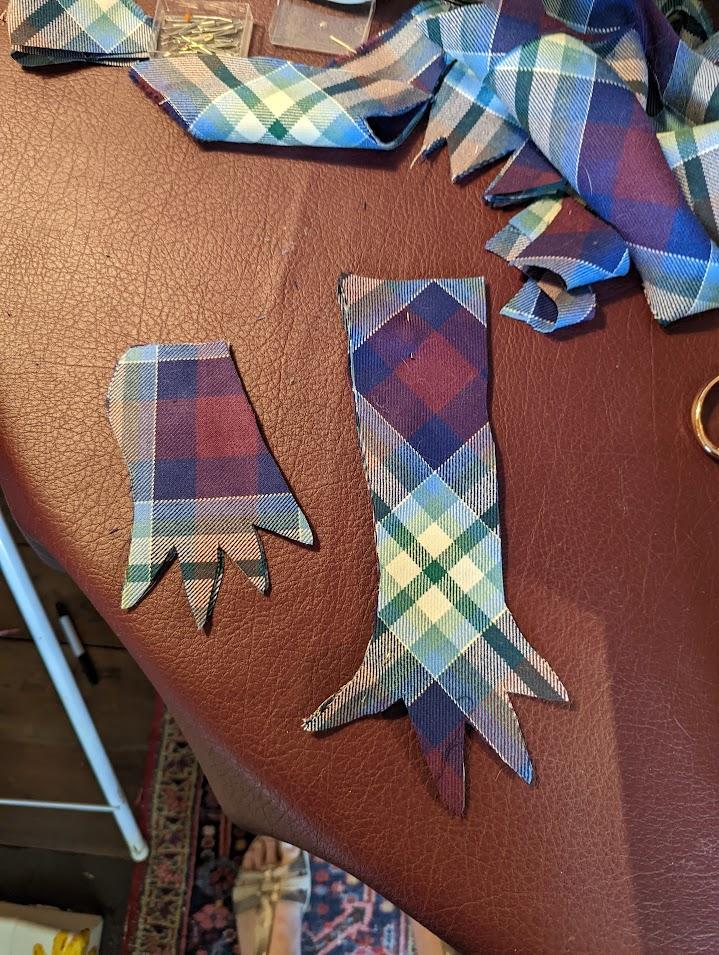 Two cutouts of tartan fabric in the shape of gloves sit atop a red worktable with fabric scraps and pins cast to the side.