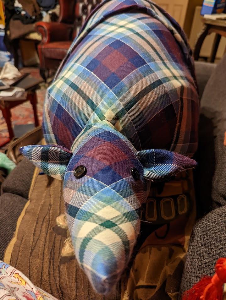 A stuffed armadillo toy made of purple, green, blue, cream, and silver Landing Zone Glasgow tartan fabric is pictured facing forward.