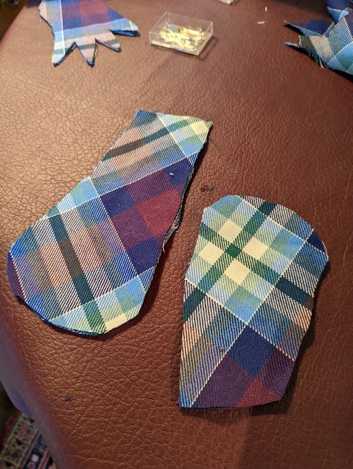 Two cutouts of tartan fabric in the shape of mittens sit on a red worktable. A cutout of a glove shape and pins are in the background.
