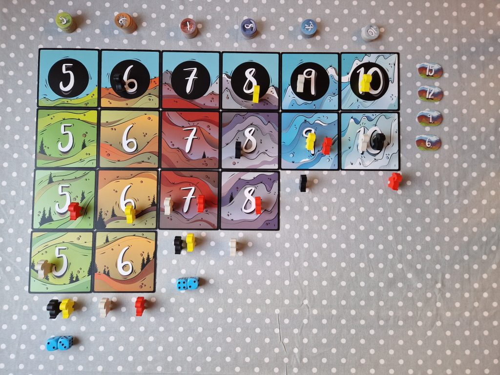 Image shows the Mountain Goats game board which includes four rows of stacked numbered squares. The squares are illustrated with rainbow-hued details of mountains. The first row has squares numbered 5 to 10, the next 5 to 8, and the last 5 and 6. There are coloured mountain goat moving pieces placed on and beside the board. Six stacks of round pieces with numbers (5-10) sit above the squares. Along the right side of the board are oval markers that number 15, 12, 9, and 6.