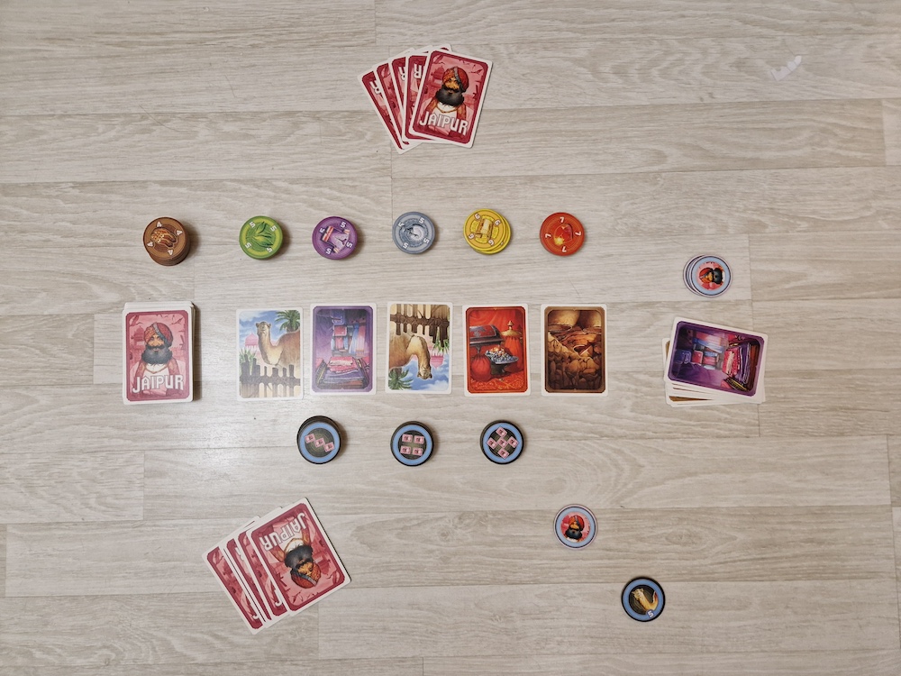 Jaipur game pieces are shown on a wood-paneled floor. Twelve piles of game tokens in various colors are shown framing a row of five illustrated cards, a draw pile, and a discard pile. To the top and bottom are two hands of four cards each. These cards are red with an illustration of a bearded man wearing a turban-style head wrap and read 'Jaipur'.