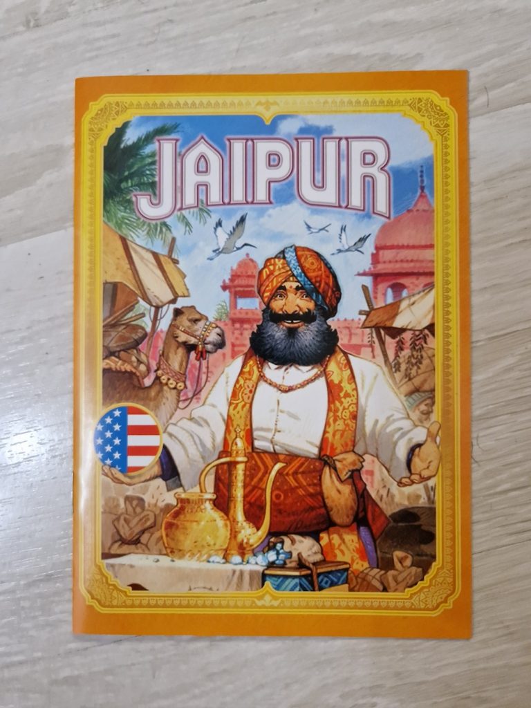 A top-down view of a board game box on a wood paneled floor. The box has border of orange and gold and an illustration of a man in a turban-style head wrap gesturing to a table full of pouches and golden wares. The background of the illustration shows a camel, three birds, and domed red buildings. 