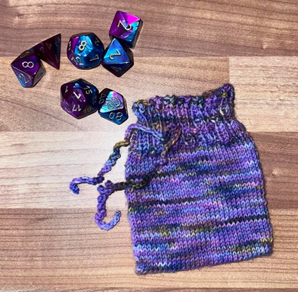 A knitted dice bag, in the Alba Aether colourway of dark and light purple, amber, blue, and black is laid flat next to seven purple and blue dice with numbers in silver. 