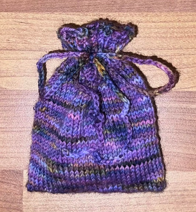 A knitted dice bag, in the Alba Aether colourway of dark and light purple, amber, blue, and black is shown. The top of the bag is closed by a bow of crocheted yarn. 