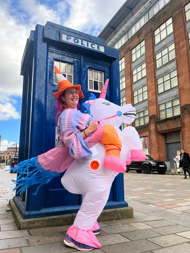 Worldcon chair, Esther MacCallum-Stewart, donning a inflatable unicorn costume, smiling and looking towards the camera, standing in front of a blue police box.