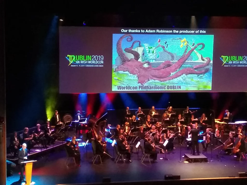 An orchestra playing on stage in front of a screen displaying an octopus conducting music and text that reads "Worldcon Philharmonic DUBLIN."