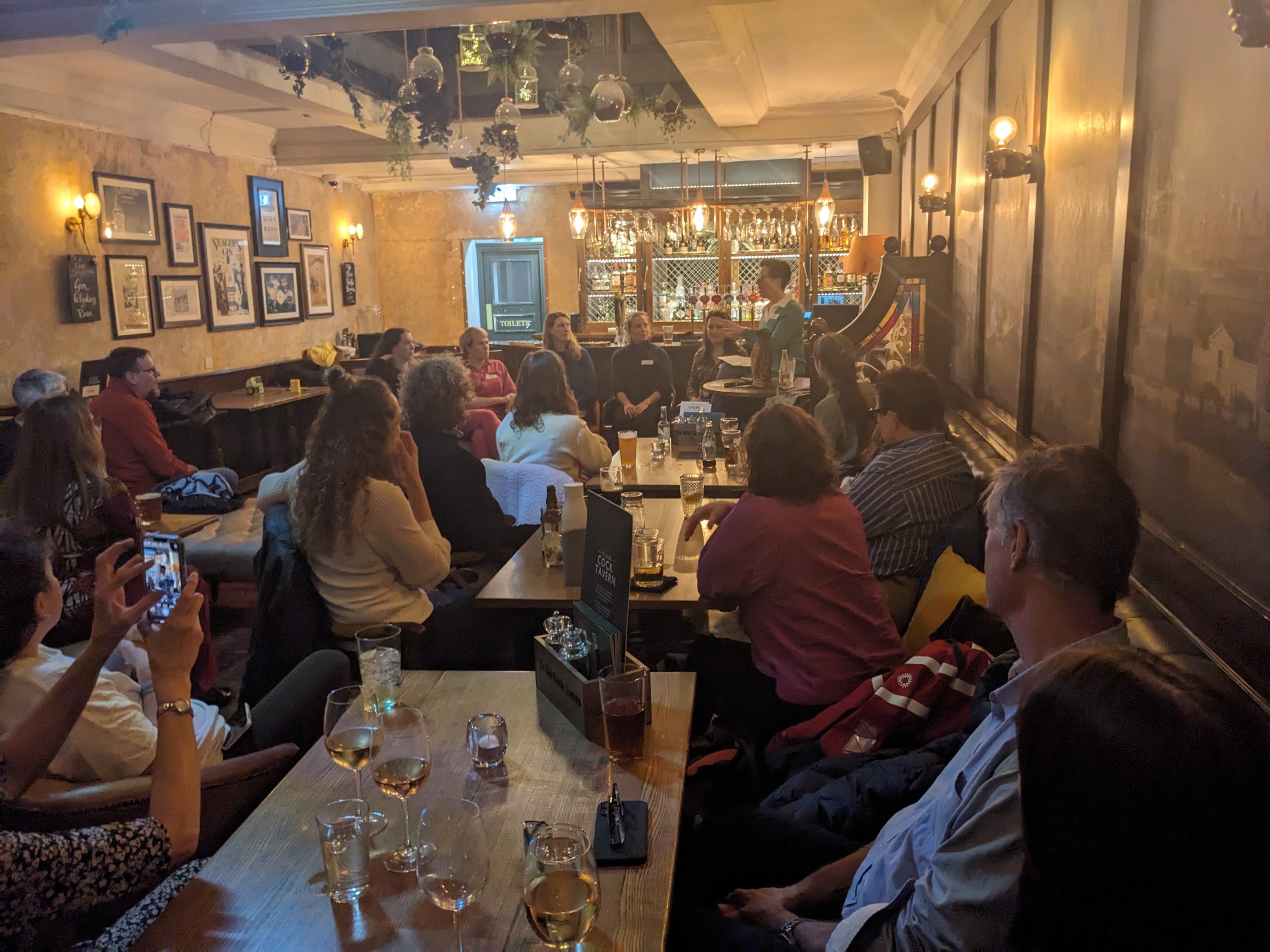 A group gathered informally around tables in a pub, watching a panel discussion.  The majority of the people in the photograph are women. 
