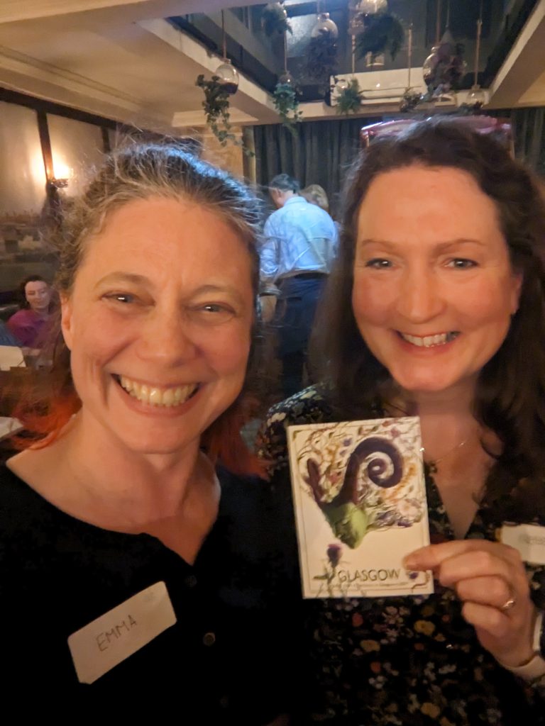 Two white women with shoulder-length brown hair smile for the
camera, holding between them a postcard showing an artistic depiction of
a fantastical woman with green skin and horns smelling a purple thistle.
The postcard says "Glasgow" at the bottom.