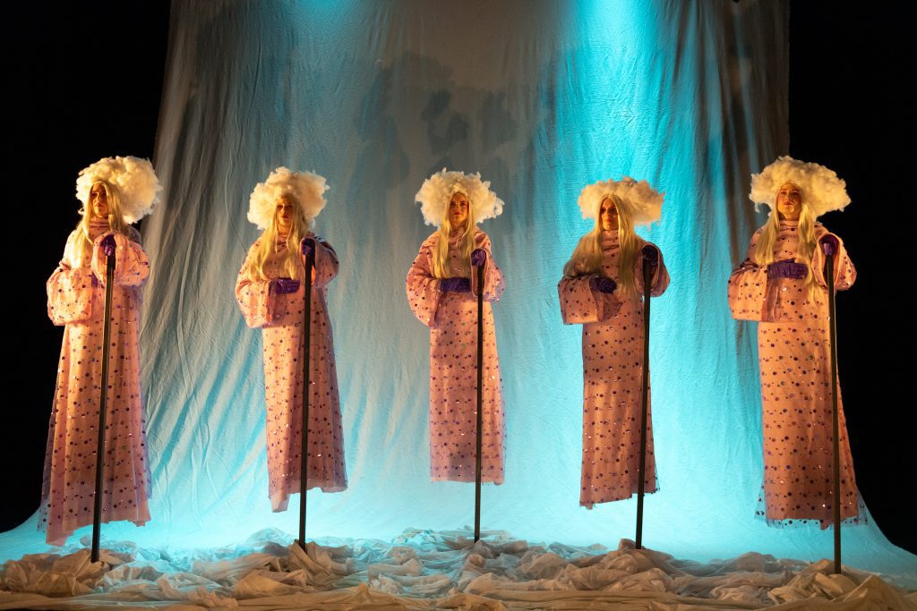 A group of five performers in colorful costumes, adorning white wigs, appear to be levitating and holding on to tall poles.